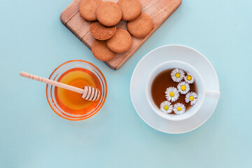 A cup of tea and honey in glass bowl with wooden dipper and cookies on cutting board on blue background. Healthy breakfast. Top view, flat lay