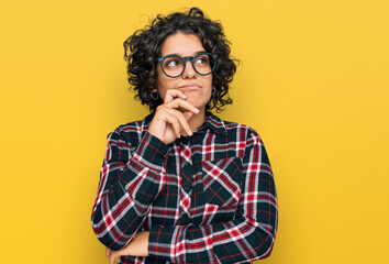 Fototapeta na wymiar Young hispanic woman with curly hair wearing casual clothes and glasses serious face thinking about question with hand on chin, thoughtful about confusing idea