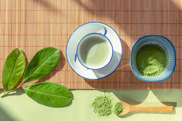 Green matcha tea in a white cup, tea powder and wooden spoon on bamboo napkin, top view. Healthy...