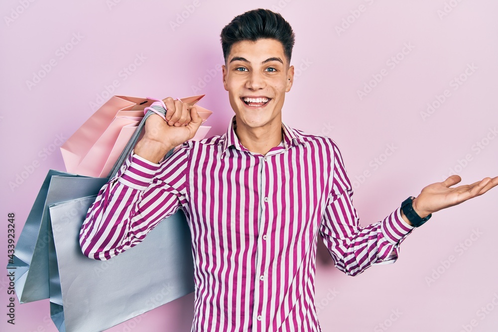 Wall mural young hispanic man holding shopping bags celebrating achievement with happy smile and winner express