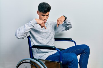 Young hispanic man sitting on wheelchair pointing down with fingers showing advertisement, surprised face and open mouth