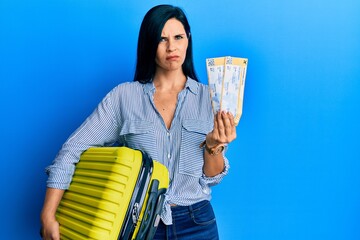 Young caucasian woman holding cabin suitcase and boarding tickets skeptic and nervous, frowning...