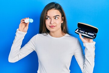 Young caucasian girl holding glasses and contact lenses smiling looking to the side and staring...