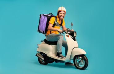 Obraz na płótnie Canvas Young courier, delivery man in uniform with thermo backpack on a moped isolated on blue background. Fast transport express home delivery. Online order.
