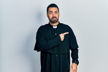 Obraz premium Handsome hispanic man with beard wearing catholic priest robe pointing aside worried and nervous with forefinger, concerned and surprised expression