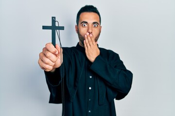Handsome hispanic priest man with beard holding catholic cross covering mouth with hand, shocked...