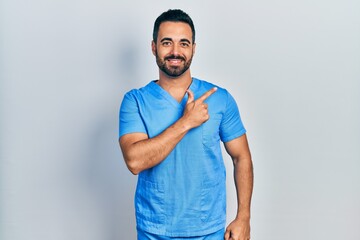 Handsome hispanic man with beard wearing blue male nurse uniform cheerful with a smile of face pointing with hand and finger up to the side with happy and natural expression on face - Powered by Adobe