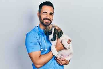 Handsome hispanic veterinary man with beard checking dog health winking looking at the camera with...