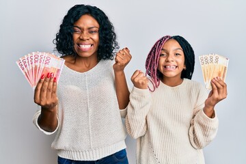 Beautiful african american mother and daughter holding norwegian krone banknotes screaming proud,...