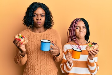 Beautiful african american mother and daughter holding sweet pastries and drinking coffee skeptic...