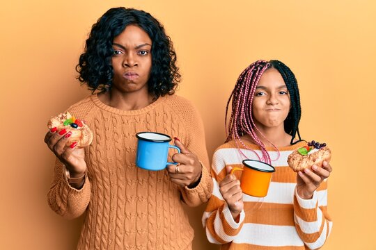Beautiful african american mother and daughter holding sweet pastries and drinking coffee skeptic and nervous, frowning upset because of problem. negative person.