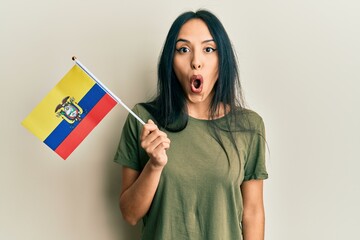 Young hispanic girl holding ecuador flag scared and amazed with open mouth for surprise, disbelief...