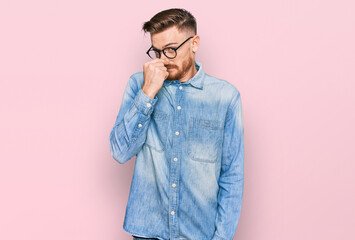 Young redhead man wearing casual denim shirt smelling something stinky and disgusting, intolerable smell, holding breath with fingers on nose. bad smell