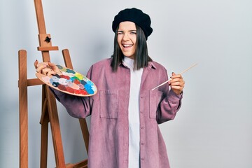 Young brunette woman standing by empty easel stand holding palette winking looking at the camera...