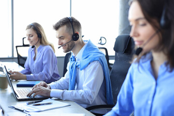 Smiling male call-center operator with headphones sitting at modern office with collegues on the...