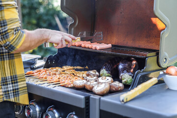 Close up shot of an anonymous man preparing meat with tongs on a barbecue outdoors. Hand of young man grilling some skewers, meat steak, sausage and vegetables on a gas grill, bbq at garden. - obrazy, fototapety, plakaty