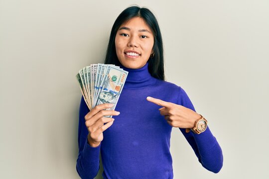 Young chinese woman holding dollars smiling happy pointing with hand and finger