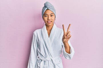 Young chinese woman wearing shower towel cap and bathrobe showing and pointing up with fingers number two while smiling confident and happy.