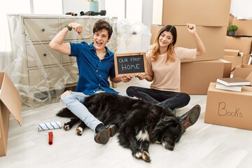 Young caucasian couple with dog holding our first home blackboard at new house strong person showing arm muscle, confident and proud of power