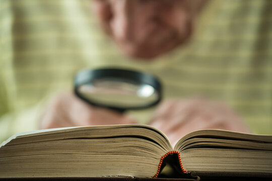 Elderly man with wrinkled hands holds magnifying glass and reads book