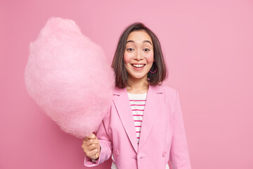 Young cheerful young woman with eastern appearance holds big candy floss smiles joyfully being in good mood dressed in fashionable clothes Positive Asian female holds sugary dessert on stick