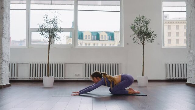 Yogi woman in sportswear doing exercises on mat in studio against windows, arching back and relaxing in Child pose. Side view flexible female practicing yoga. Concept of fitness