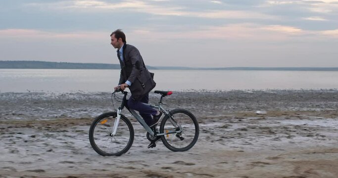 A businessman quickly rides a bicycle along the coast of the lake, overcoming a difficult sandy road against the backdrop of a cloudy sky.