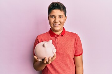 Fototapeta na wymiar Teenager hispanic boy holding piggy bank looking positive and happy standing and smiling with a confident smile showing teeth