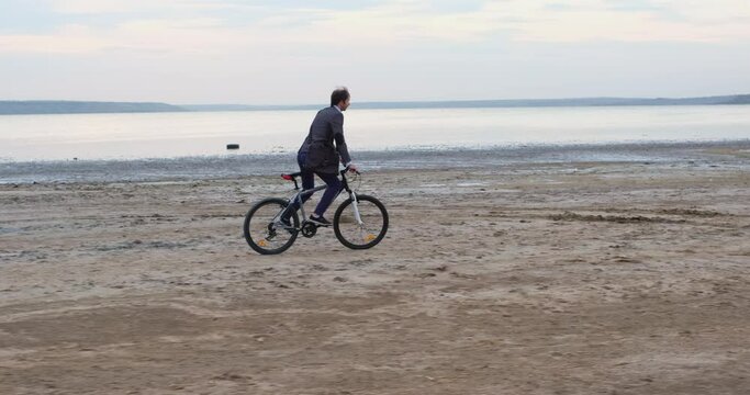A business man on a bicycle overcomes a difficult section of the path, moving along the coast against the backdrop of a cloudy sunset.