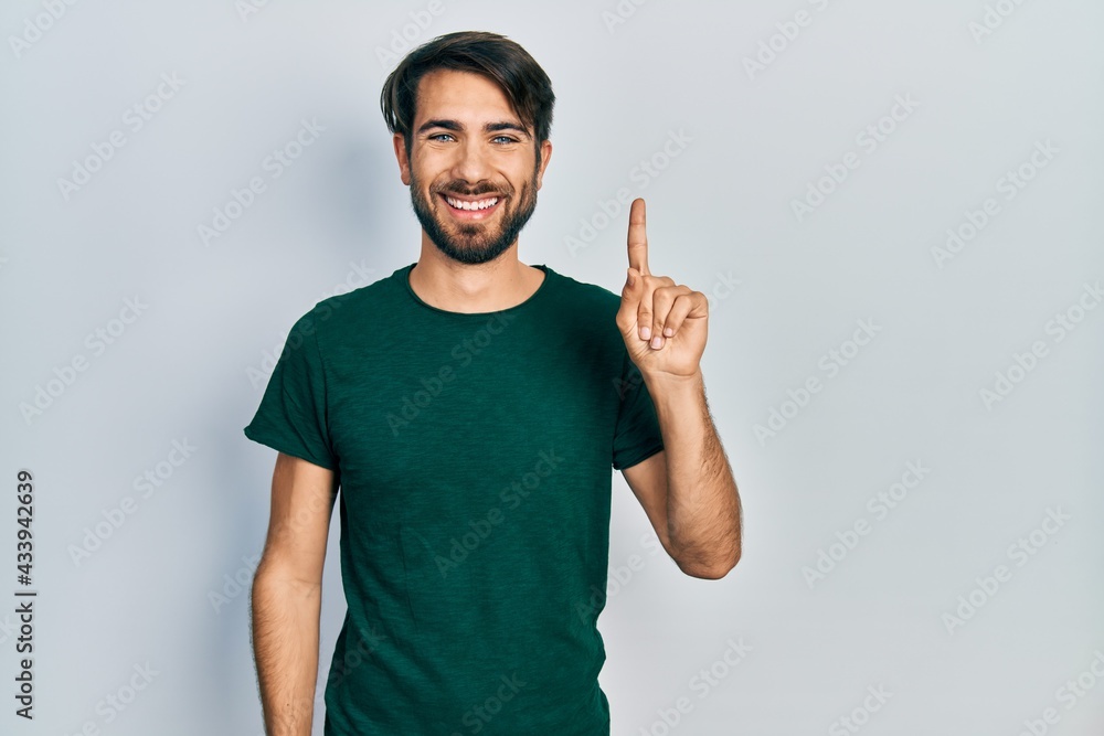 Wall mural Young hispanic man wearing casual white tshirt showing and pointing up with finger number one while smiling confident and happy.