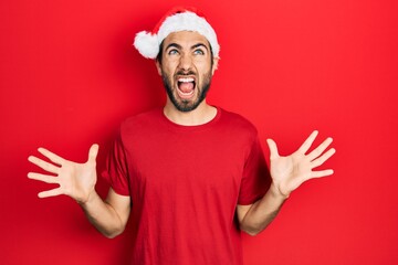 Young hispanic man wearing christmas hat crazy and mad shouting and yelling with aggressive expression and arms raised. frustration concept.