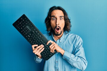 Young hispanic man holding keyboard afraid and shocked with surprise and amazed expression, fear...