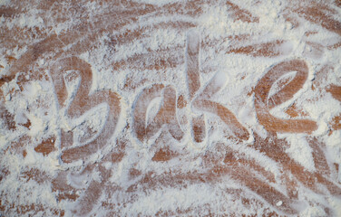The word Bake written into messily scattered flour revealing wooden board below. The concept of home-made bread baking.