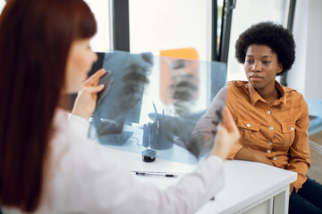 View from the female doctor shoulder, holding x-ray chest scan, explaining the results to patient. Focus on young african woman listening to the doctor recommendations