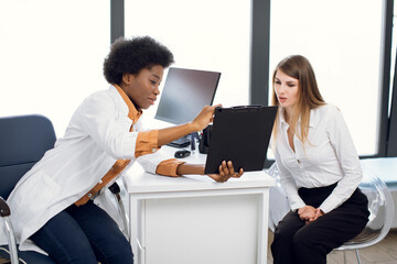 Fototapeta premium African American female doctor and pretty Caucasian woman patient Discussing the way of treatment or medical record data at modern diagnostic center. Doctor shows a clipboard to patient