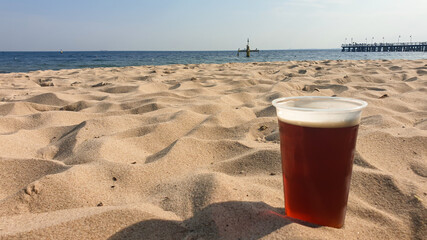 A beer in a plastic cup located on the sandy beach in Gdynia, Poland. In the back there is a small...
