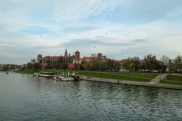 Obraz premium A panoramic view on the iconic Wawel Castle in Cracow, Poland and the Vistula river flowing under the castle. A few birds flying above the river. A few boats docked on the rivers bend. City tour.