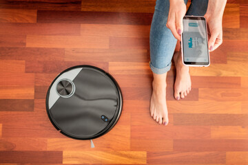 Young girl feet at home using a smart phone to control a round smart automatic vacuum, machine...