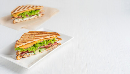 Homemade juicy sandwiches made of cucumber, meat and cheese, lettuce between slices of grilled toast bread served on plate and on paper on white wooden background at kitchen. Image with copy space