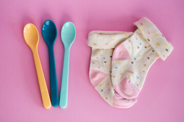 Baby plastic spoons and cute stylish socks in pastel colours. Baby accessories on pink background, top view. Cutlery for children. 
