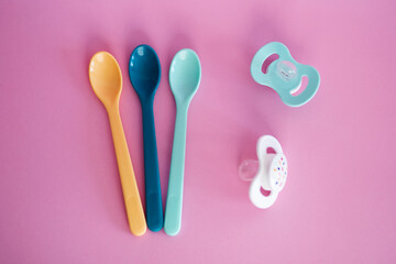 Baby plastic spoons and pacifiers in pastel colours. Baby accessories on pink background, top view. Cutlery for children. 