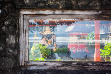 Defocused anonymous girl in a straw hat and dress reflected in a window pane showing bright Balinese temple and garden
