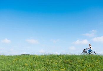 woman rides bicycle on grassy dike with flowers under blue sky