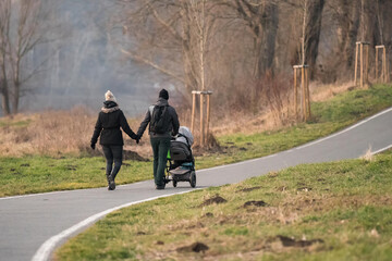 Couple with a baby-carriage walking in spring forest on buildings background in the distance. Young parents with pram, concept of eco-friendly residential area