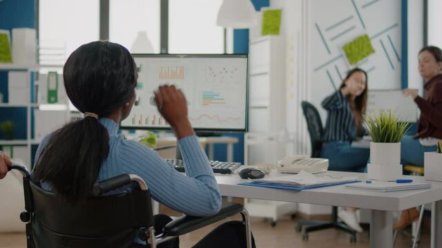 Man employee helping his invalid african colleague coming in workplace pushing wheelchair near desk. Black handicapped paralysed woman greetting colleagues starting working at financial project