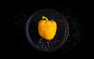 Fototapeta na wymiar Yellow pepper with a green stalk falls into the water in a black bowl on a black background.