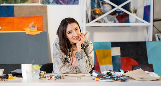 Drawing class. Art school. Creative occupation. Talented happy smiling left-handed female artist sitting with pencil at messy workplace with painting supplies in modern workshop.