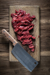 fresh raw meat cut into pieces for cooking beef stroganoff lies on a wooden cutting board with a kitchen cleaver on a background of old veneer