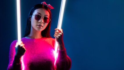 Neon light beauty. Cyberpunk girl. Futuristic fashion. Asian woman in sunglasses in glamour pink red color glow on white LED swing isolated on dark night blue copy space background.