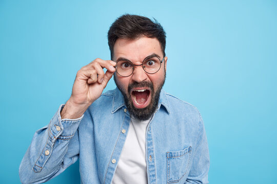 Furious bearded annoyed adut man screams loudly yells upset keeps hand on rim of spectacles expresses irritation dressed in denim shirt isolated over blue background has argument with somone